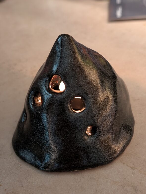 A clay candle holder, black with holes. Through the holes is a flame.
